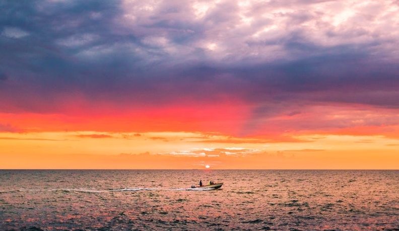 Boat on the ocean with an orange sunset and dramatic sky. Photo by Mantas Hesthaven on Pexels.com